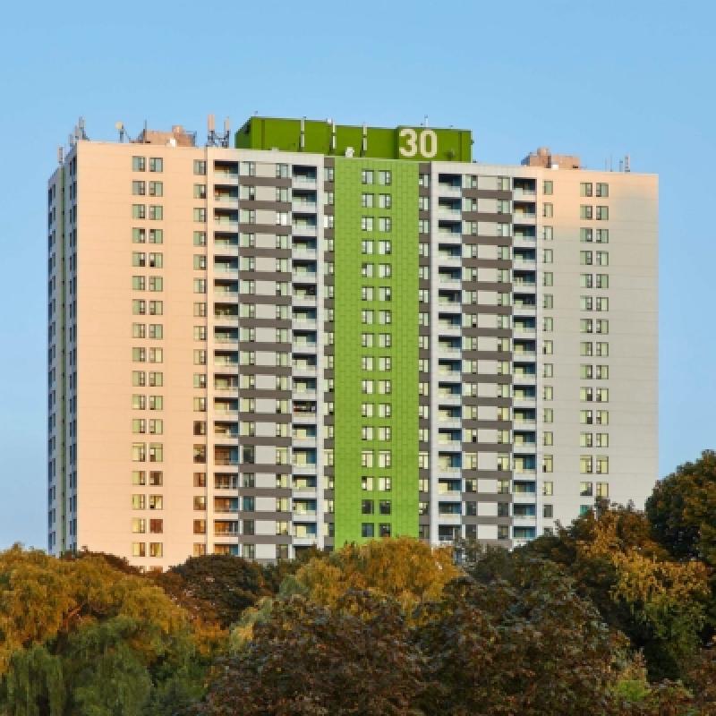 Tall apartment building with a green central stripe and the number "30" on top. Surrounding trees and clear blue sky create a serene urban scene.